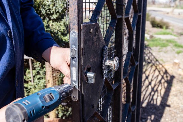 Worker fixing a door lock on an iron wicket with an electric screwdriver, showcasing door repair work outdoors, essential for security maintenance.